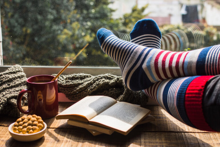 A pair of feet in long winter socks on a windowsill, there is a cup of coffee, a little bowl of snacks and books on the windowsill too.