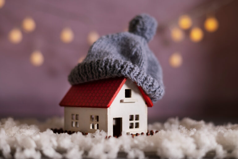A small toy house with a small beanie on the roof surrounded by cotton balls that look like snow.