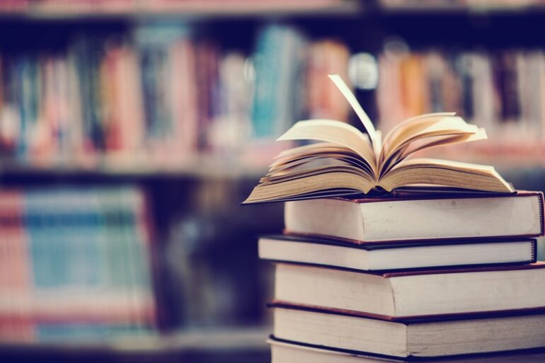 An open book on top of a stack of books. a blurred view of library shelves can be seen in the background.
